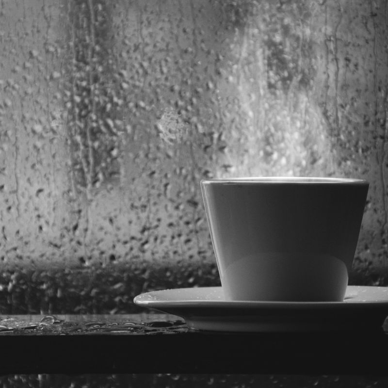 stock photo of cup by window during rain a white cup with steam coming out of the top on a white plate in front of a window that shows it's raining outside